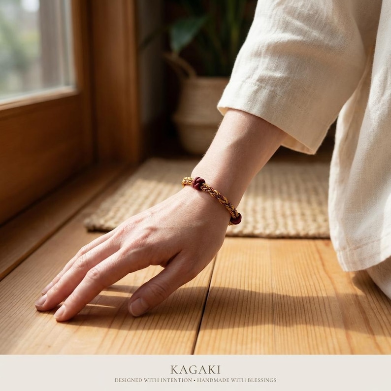 May include: A gold-toned bracelet with woven detailing and dark red beads. The bracelet is worn on a wrist, with the hand resting on a wooden surface. The word "KAGAKI" is printed at the bottom of the image.
