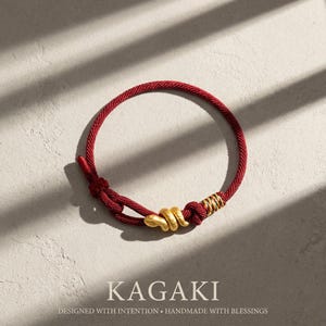 May include: A red cord bracelet with a gold-colored snake-shaped charm and gold-colored accents. The bracelet is knotted and features the text "KAGAKI" and "DESIGNED WITH INTENTION • HANDMADE WITH BLESSINGS".