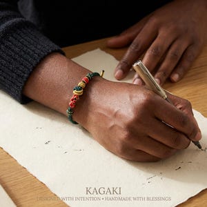 May include: A green, red, and gold beaded bracelet is worn on a wrist. The bracelet is made of knotted cord and features decorative beads. The image includes the text "KAGAKI" and "DESIGNED WITH INTENTION &bull; HANDMADE WITH BLESSINGS".