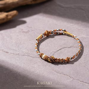 May include: A braided bracelet with a mix of brown, white, and green threads. The bracelet features a central decorative element with a floral design. The text "KAGAKI" and "DESIGNED WITH INTENTION &bull; HANDMADE WITH INTENTION" are visible.