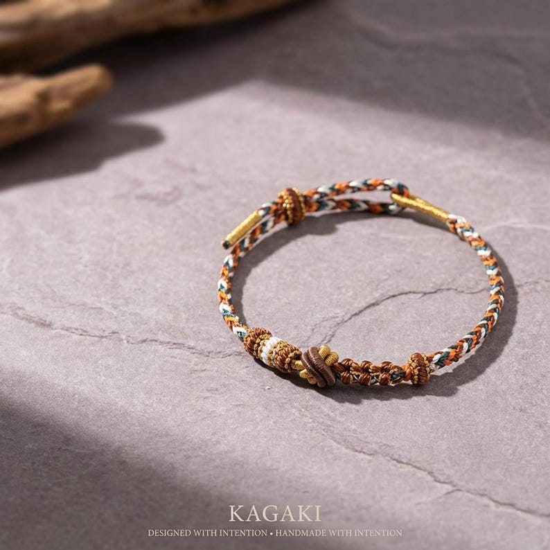 May include: A braided bracelet with a mix of brown, white, and green threads. The bracelet features a central decorative element with a floral design. The text "KAGAKI" and "DESIGNED WITH INTENTION &bull; HANDMADE WITH INTENTION" are visible.