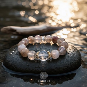 May include: A bracelet featuring a central clear bead and smaller, speckled beads in shades of tan, brown, and clear. The bracelet sits on a dark, smooth stone, with water and sunlight in the background. The brand name "KAGAKI" is visible.
