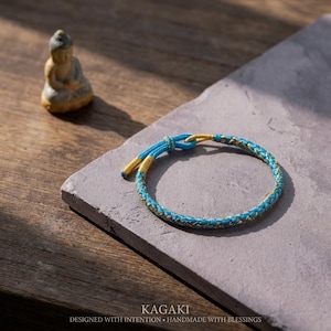 May include: A turquoise and gold braided bracelet with a knot closure. The bracelet is on a gray stone surface. The text "KAGAKI" is at the bottom, with the words "DESIGNED WITH INTENTION • HANDMADE WITH BLESSINGS" below it.