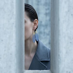 May include: A person wearing blue feather earrings with yellow accents. The earrings are suspended from silver hooks. The person is wearing a gray coat. The background is a light blue.