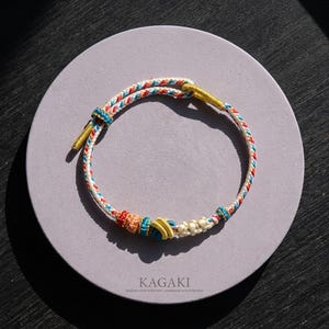 May include: A handmade bracelet with a braided design in white, red, blue, and yellow. The bracelet is knotted with decorative elements and gold-colored accents. The text "KAGAKI" is visible on the surface below the bracelet.