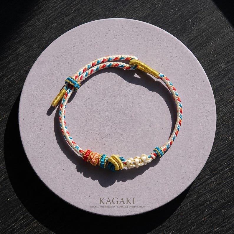 May include: A handmade bracelet with a braided design in white, red, blue, and yellow. The bracelet is knotted with decorative elements and gold-colored accents. The text "KAGAKI" is visible on the surface below the bracelet.