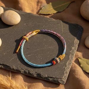 May include: A colorful braided bracelet with blue, pink, and gold accents. The bracelet is made of woven threads and features a decorative knot. The bracelet is displayed on a dark gray stone surface.