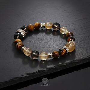 Könnte beinhalten: Ein Armband mit einer Mischung aus braunen, goldenen und klaren Edelsteinen. Das Armband enthält Tigerauge-, Rauchquarz- und Rutilquarz-Perlen mit silberfarbenen Abstandshaltern und dekorativen Akzenten. Das Armband wird auf einer dunklen Oberfläche präsentiert.