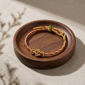 May include: A gold-colored bracelet with a knot design, resting on a wooden plate. The bracelet features red, yellow, blue, and green threads. The wooden plate is dark brown. The bracelet is a piece of jewelry.