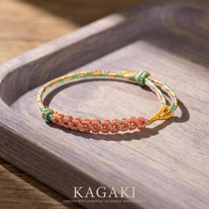 May include: A handmade bracelet with a floral design. The bracelet features a series of small, orange-red flower-shaped knots. The cord is multi-colored, with green, yellow, and pink hues. The word "KAGAKI" is printed at the bottom, along with the phrase "DESIGNED WITH INTENTION • HANDMADE WITH BLESSINGS."