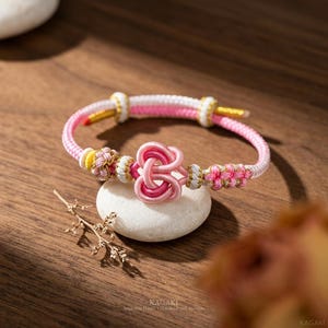 Könnte beinhalten: Ein rosa-weiß geflochtenes Armband mit einem zentralen Knotendesign und floralen Akzenten. Das Armband wird auf einem weißen Stein vor dunklem Holz präsentiert. Goldfarbene Details verleihen dem handgefertigten Accessoire Eleganz.