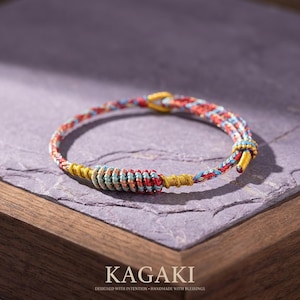 May include: A colorful handmade bracelet with a woven design. The bracelet features red, blue, and yellow threads, with a gold-colored clasp. The word "KAGAKI" is printed at the bottom of the image, along with the text "DESIGNED WITH INTENTION • HANDMADE WITH BLESSINGS."