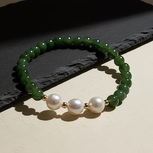 May include: A beaded bracelet featuring dark green jade beads and three large, round white pearls. The pearls are separated by small gold-colored beads. The bracelet is displayed on a dark gray surface.