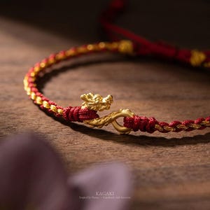 May include: A bracelet featuring a braided red and gold cord with a gold-colored dragon-shaped clasp. The clasp consists of a dragon head and a tail that hook together. The text KAGAKI Inspired by Nature Handmade with Intention is visible.