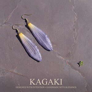 May include: A pair of handmade earrings featuring elongated, lavender-colored petals with yellow tips, suspended from gold-colored hooks. The earrings are set against a gray background. The text "KAGAKI" and "DESIGNED WITH INTENTION &bull; HANDMADE WITH BLESSINGS" are at the bottom.
