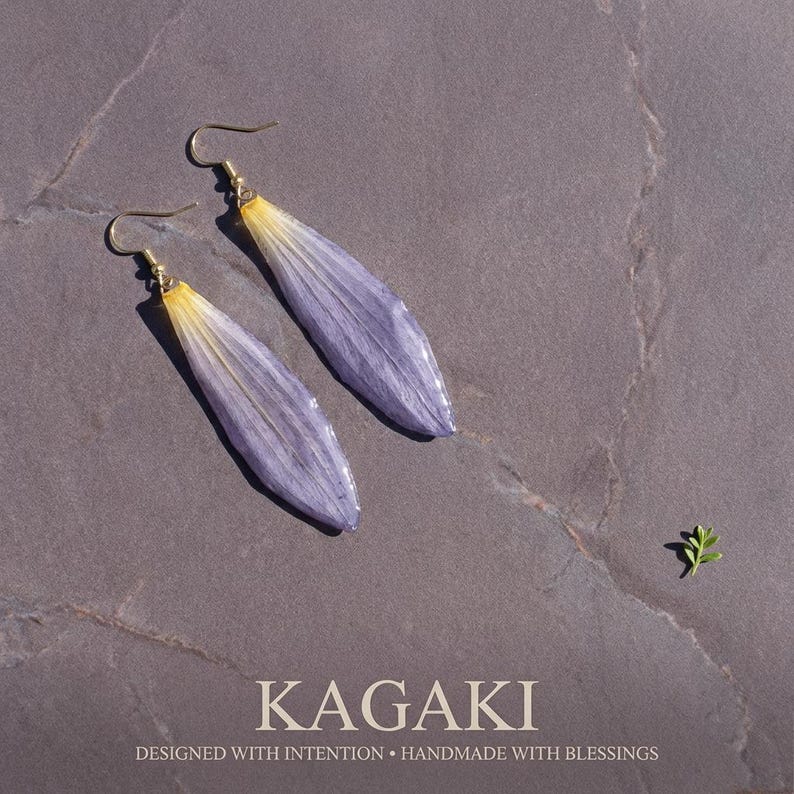 May include: A pair of handmade earrings featuring elongated, lavender-colored petals with yellow tips, suspended from gold-colored hooks. The earrings are set against a gray background. The text "KAGAKI" and "DESIGNED WITH INTENTION &bull; HANDMADE WITH BLESSINGS" are at the bottom.
