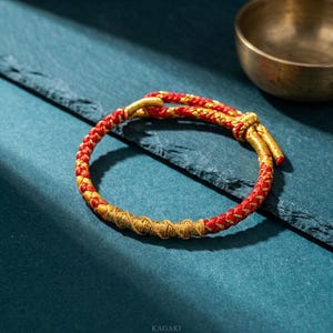 May include: A red and gold braided bracelet with a knot closure. The bracelet has a woven design with alternating red and gold threads. The bracelet is on a dark blue surface. The word "KAGAKI" is at the bottom of the image.