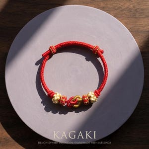 May include: A red braided bracelet with gold-colored accents and decorative knots. The bracelet is displayed on a gray circular surface. The text "KAGAKI" and "DESIGNED WITH INTENTION + HANDMADE WITH BLESSINGS" are at the bottom.