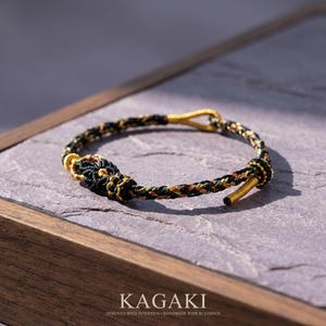May include: A braided bracelet with black, gold, and orange threads. The bracelet has gold-colored metal accents and a clasp. The word "KAGAKI" is at the bottom of the image, along with the text "DESIGNED WITH INTENTION • HANDMADE WITH BLESSINGS."