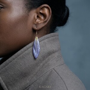 May include: A close-up of a gold-toned earring with a long, teardrop-shaped pendant. The pendant is a pale purple color with a yellow tip, resembling a flower petal. The earring is worn on a person's ear. The word "KAGAKI" is at the bottom.