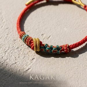 May include: Close-up of a red braided bracelet with gold and teal accents. The bracelet features intricate knotwork and floral details. The word "KAGAKI" is displayed in a serif font, with the text "DESIGNED WITH INTENTION &bull; HANDMADE WITH INTENTION" below.