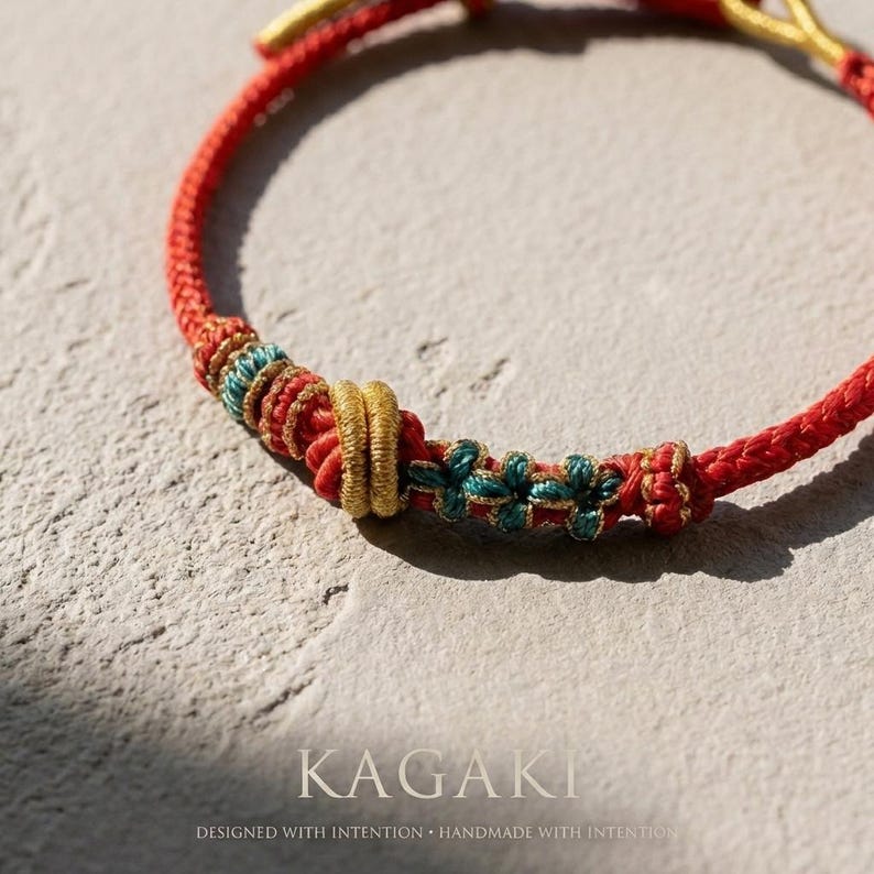 May include: Close-up of a red braided bracelet with gold and teal accents. The bracelet features intricate knotwork and floral details. The word "KAGAKI" is displayed in a serif font, with the text "DESIGNED WITH INTENTION &bull; HANDMADE WITH INTENTION" below.