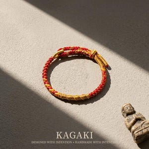 May include: A red and gold braided bracelet with a knot closure. The bracelet is on a neutral surface with the text "KAGAKI" and "DESIGNED WITH INTENTION &bull; HANDMADE WITH INTENTION" below it. A small statue is in the background.