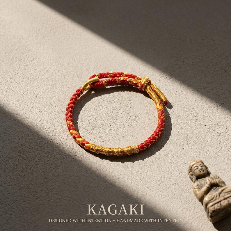 May include: A red and gold braided bracelet with a knot closure. The bracelet is on a neutral surface with the text "KAGAKI" and "DESIGNED WITH INTENTION &bull; HANDMADE WITH INTENTION" below it. A small statue is in the background.