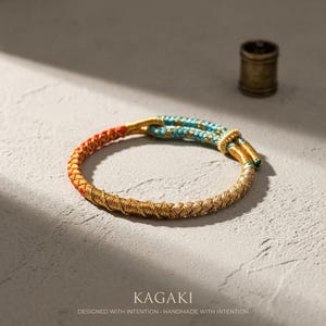 May include: A braided bracelet with gold, red, and turquoise accents. The bracelet features a woven design with a gold base, transitioning to red and turquoise sections. The word "KAGAKI" is displayed below the bracelet.