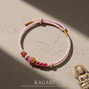 May include: A pink and gold bracelet with intricate knotwork and decorative accents. The bracelet features a braided cord and gold-colored metal details. The text "KAGAKI" and "DESIGNED WITH INTENTION &bull; HANDMADE WITH INTENTION" are at the bottom of the image.