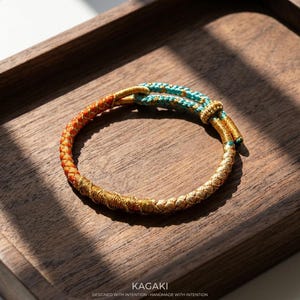 May include: A handcrafted bracelet with a braided design. The bracelet features a color gradient, transitioning from orange to gold and ending with a turquoise accent. The bracelet is displayed on a wooden tray. The text "KAGAKI" is visible.