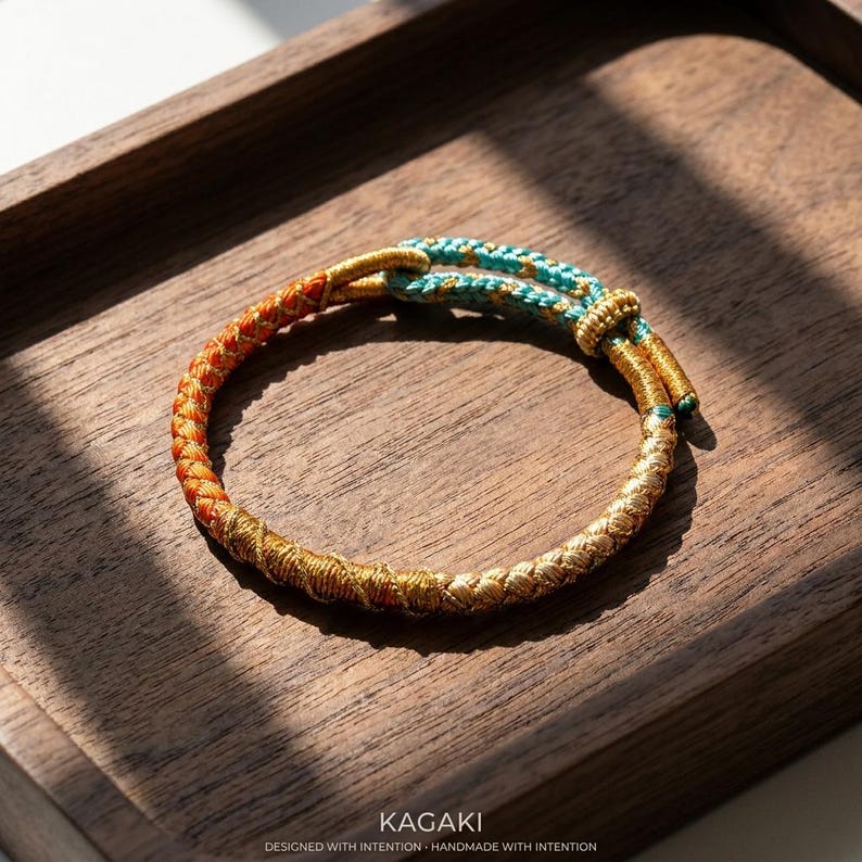 May include: A handcrafted bracelet with a braided design. The bracelet features a color gradient, transitioning from orange to gold and ending with a turquoise accent. The bracelet is displayed on a wooden tray. The text "KAGAKI" is visible.