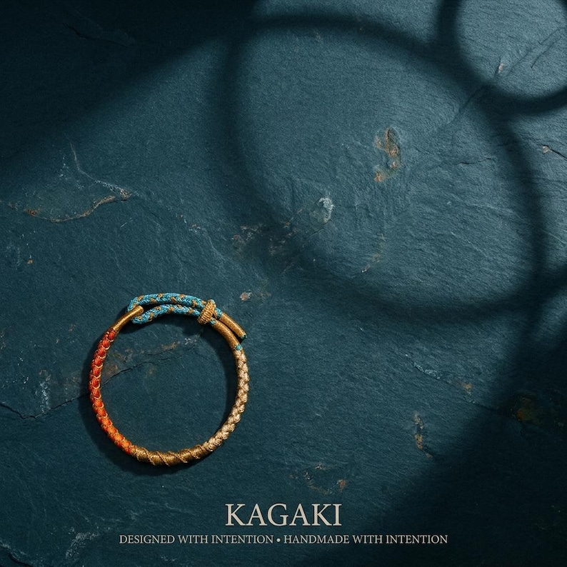 May include: A handcrafted bracelet with a braided design. The bracelet features a gradient of colors, transitioning from orange to gold, with a turquoise knot. The word "KAGAKI" is displayed below the bracelet, along with the text "DESIGNED WITH INTENTION &bull; HANDMADE WITH INTENTION."