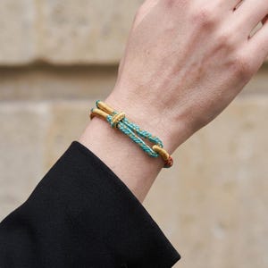 May include: A gold and turquoise rope bracelet with a knotted design. The bracelet features a gold clasp and accents, with a turquoise and gold rope. The bracelet is worn on a wrist.
