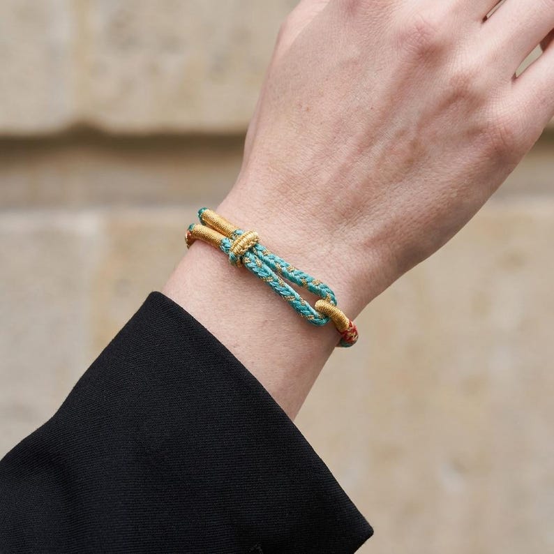May include: A gold and turquoise rope bracelet with a knotted design. The bracelet features a gold clasp and accents, with a turquoise and gold rope. The bracelet is worn on a wrist.