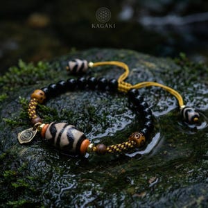 May include: A beaded bracelet with a central dzi bead, black beads, and decorative wooden and gold accents. The bracelet is set against a mossy rock background. The brand name "KAGAKI" is visible in the upper right corner.