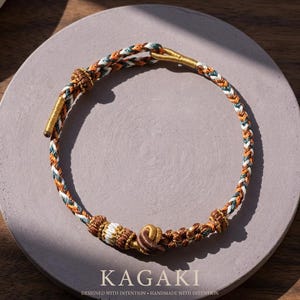 May include: Braided necklace with orange, white, and green threads, accented with gold-colored metal. The necklace is circular, with a central knot and a clasp. The word "KAGAKI" is printed at the bottom, along with the text "DESIGNED WITH INTENTION &bull; HANDMADE WITH INTENTION."