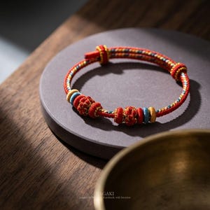 May include: A red, yellow, and blue braided bracelet with decorative knots and beads. The bracelet is displayed on a gray circular surface, with a wooden background and a gold-colored bowl in the foreground.