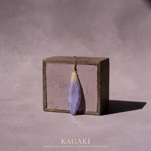 May include: A single, delicate earring featuring a lavender-colored petal suspended from a gold-colored hook. The earring is displayed against a rustic, wooden block with a lavender-colored interior. The word "KAGAKI" is at the bottom.