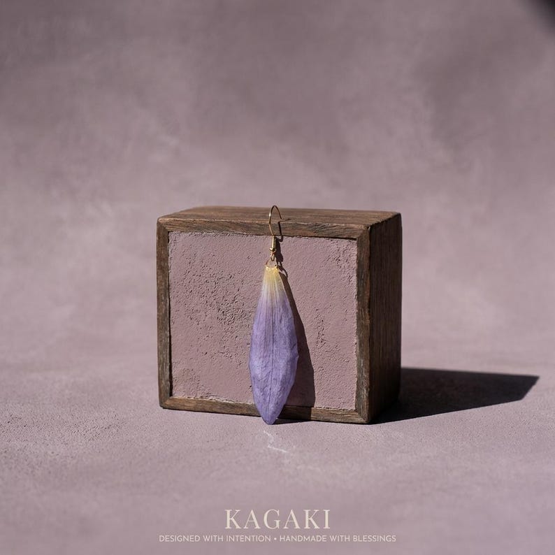 May include: A single, delicate earring featuring a lavender-colored petal suspended from a gold-colored hook. The earring is displayed against a rustic, wooden block with a lavender-colored interior. The word "KAGAKI" is at the bottom.