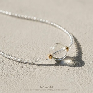 Pode incluir: Um colar de cristal de quartzo transparente com uma grande conta esférica e contas menores. O colar apresenta detalhes dourados e é apresentado em uma superfície neutra. O texto "KAGAKI" é visível na parte inferior.