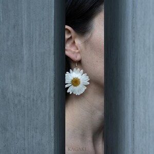 Op de afbeelding: Een close-up van een madeliefjesoorbel met witte bloemblaadjes en een gele kern. De oorbel hangt aan een goudkleurige haak. Het woord "KAGAKI" is zichtbaar onderaan de afbeelding. De oorbel wordt door een persoon gedragen.