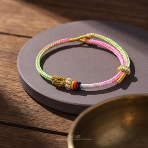 Può includere: Un braccialetto fatto a mano con un design intrecciato. Il braccialetto presenta una combinazione di fili verdi, rosa e bianchi, accentuati da nodi color oro e piccole perline colorate. Il braccialetto è presentato su una superficie grigia.