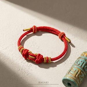 May include: Red and gold braided bracelet with intricate knot details. The bracelet features a sliding knot closure and gold accents. The word "KAGAKI" is printed at the bottom of the image.