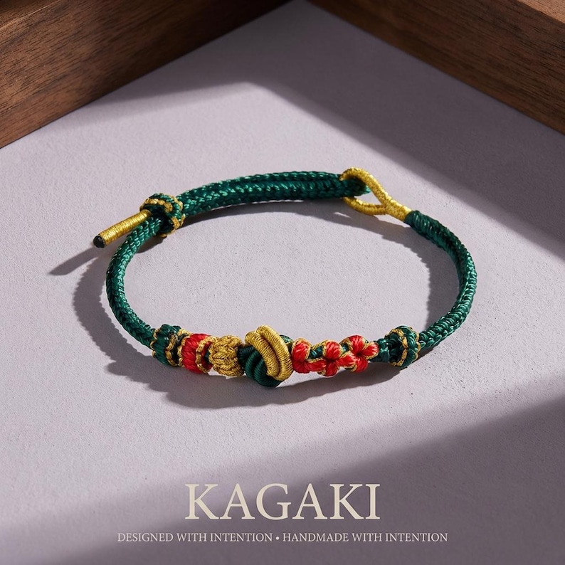 May include: Green and gold braided bracelet with red accents. The bracelet features a central knot design with red and gold details. The text "KAGAKI" is displayed at the bottom, along with the phrase "DESIGNED WITH INTENTION &bull; HANDMADE WITH INTENTION."