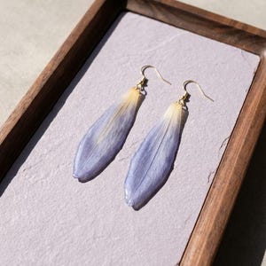 May include: A pair of dangle earrings featuring elongated, lavender-blue petals with a gradient of yellow at the top. The earrings have gold-colored hooks and are displayed on a light purple surface within a wooden tray.