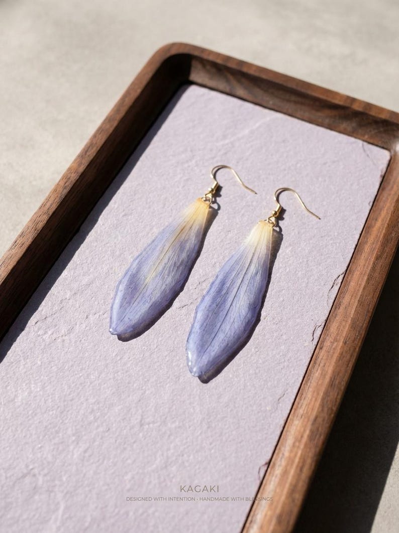 May include: A pair of dangle earrings featuring elongated, lavender-blue petals with a gradient of yellow at the top. The earrings have gold-colored hooks and are displayed on a light purple surface within a wooden tray.