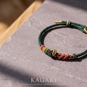 May include: A green and gold bracelet with red accents. The bracelet features intricate knotwork and decorative elements. The word "KAGAKI" is displayed at the bottom of the image, along with the text "DESIGNED WITH INTENTION &bull; HANDMADE WITH INTENTION."