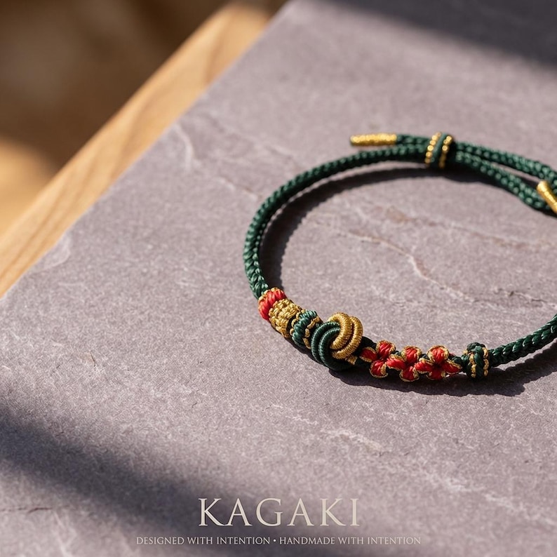 May include: A green and gold bracelet with red accents. The bracelet features intricate knotwork and decorative elements. The word "KAGAKI" is displayed at the bottom of the image, along with the text "DESIGNED WITH INTENTION &bull; HANDMADE WITH INTENTION."