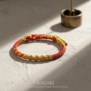 May include: A braided bracelet with red and gold threads. The bracelet has a woven design with gold accents and a clasp. The word "KAGAKI" is printed at the bottom of the image, along with the text "DESIGNED WITH INTENTION &bull; HANDMADE WITH INTENTION."